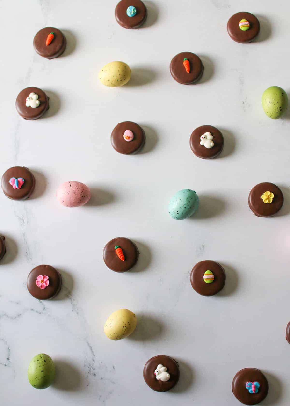 Overhead of Chocolate covered oreos with festive easter royal icing decorations on a white marble background surrounded by pastel freckled eggs.
