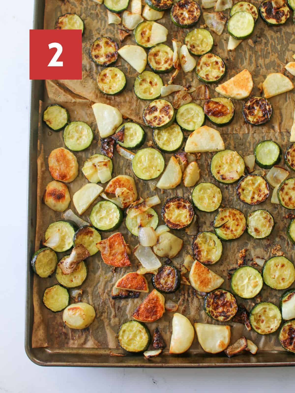 Roasted potatoes, zucchini, onion and garlic on a large sheet pan with brown parchment paper on a white marble background with a red box with a white '2' in the upper left corner.
