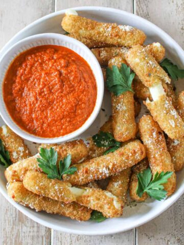 Overhead of Air Fryer Frozen Mozzarella Sticks in a grey white low bowl topped with parmesan and italian parsley. In the corner is a small bowl of marinara sauce.