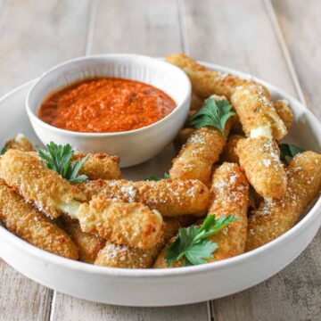 Angled photo of Air Fryer Frozen Mozzarella Sticks in a grey white low bowl topped with parmesan and italian parsley. The white grey low bowl sits on a wood plank board. In the corner is a small bowl of marinara sauce.