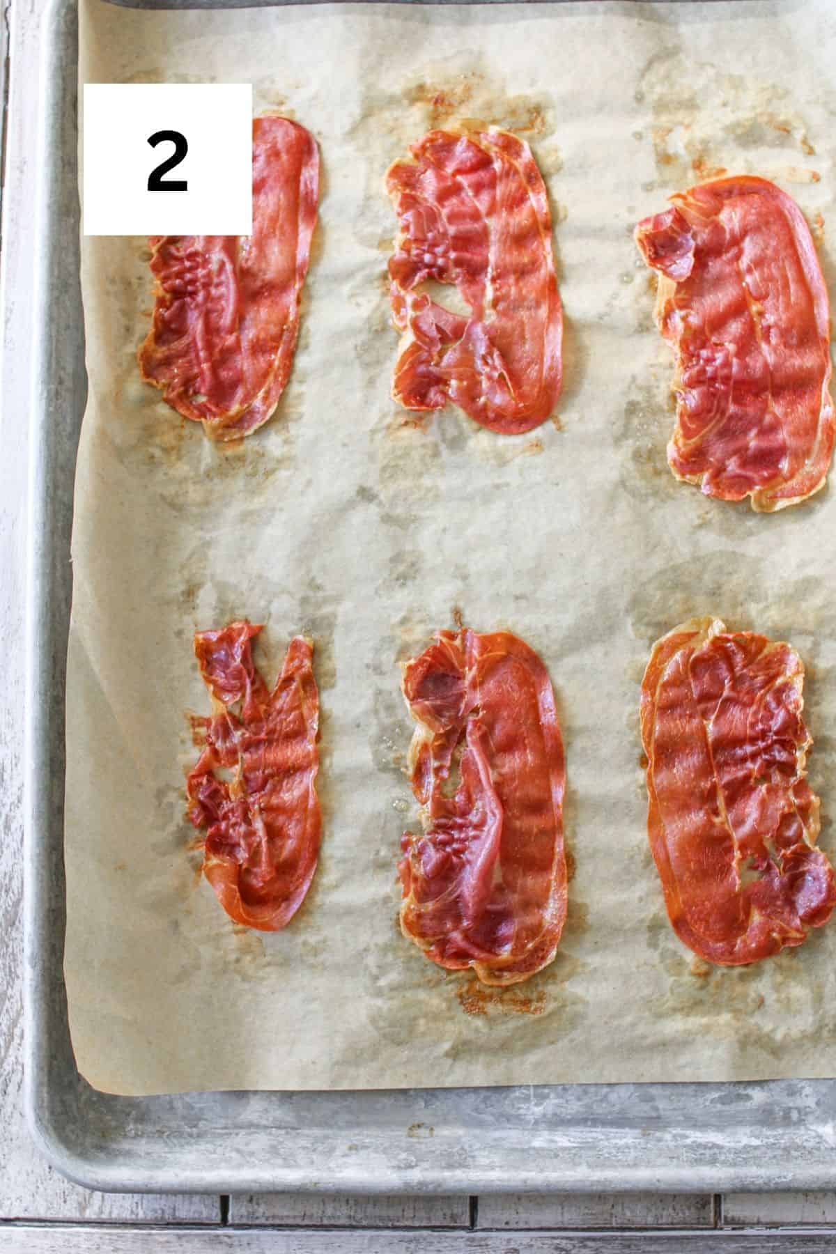6 slices of crispy prosciutto on a piece of brown parchment on a baking sheet on a light wood plank background. A white square is in the upper left corner with a black '2'.