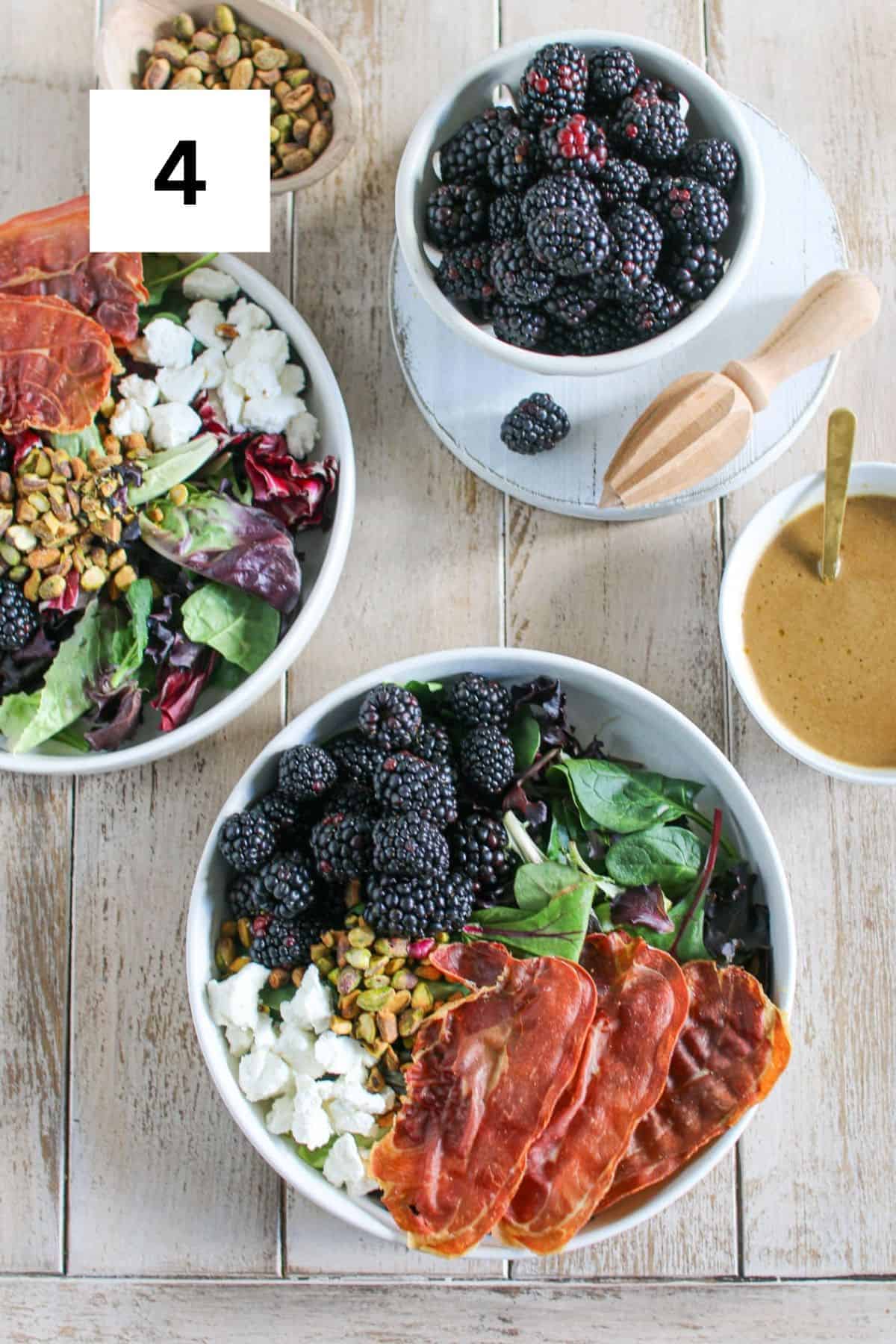 Two bowls of Blackberry Goat Cheese Salad on a wood plank background. Each bowl has a gold fork with it and theres a small bowl of blackberries, a small oval wood bowl of pistachios and a circle white bowl of dressing.A white square is in the upper left corner with a black '4'.