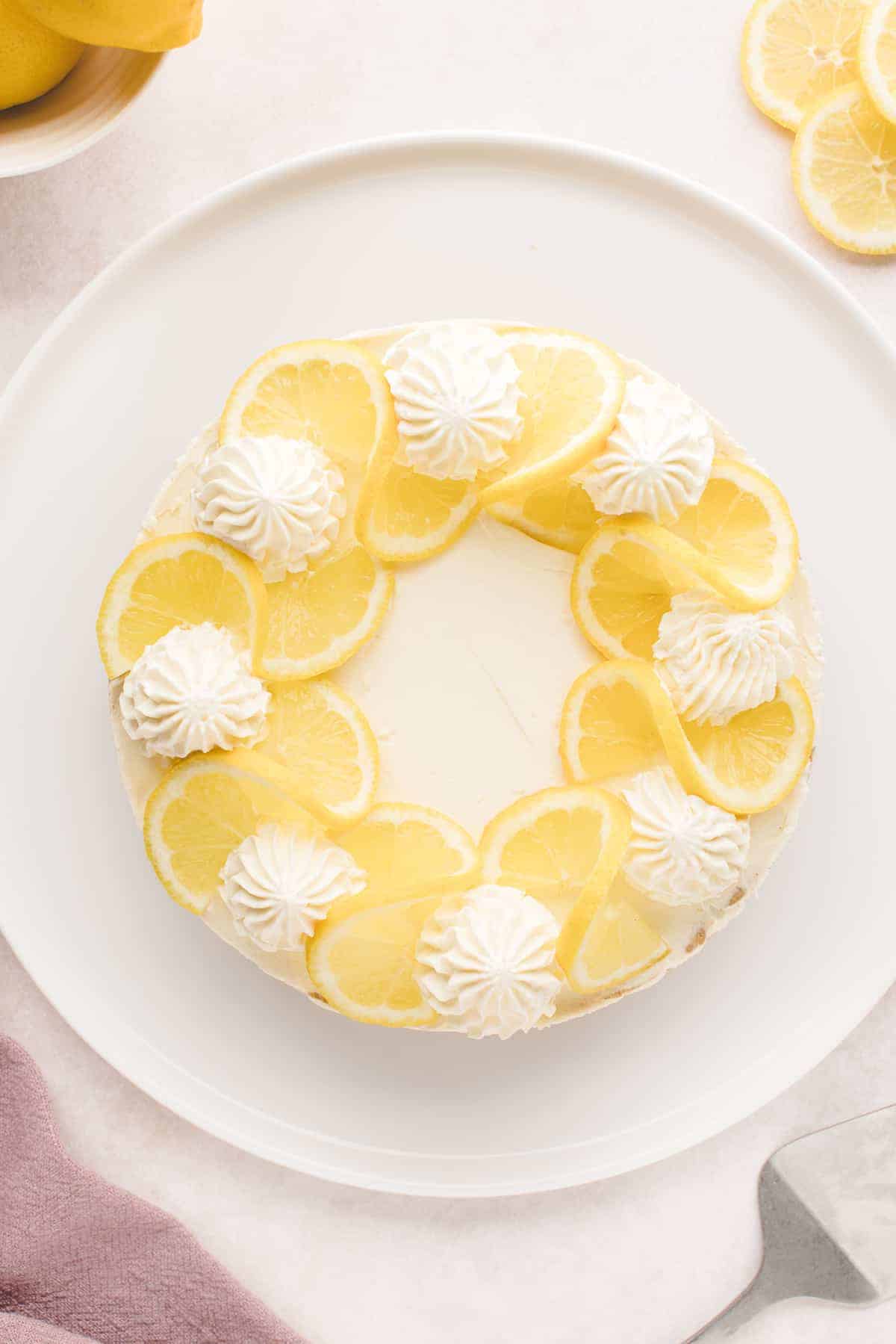 Overhead No Bake Lemon Cheesecake on a white platter. The top is decorated with piped whipped cream and twisted lemon slices.