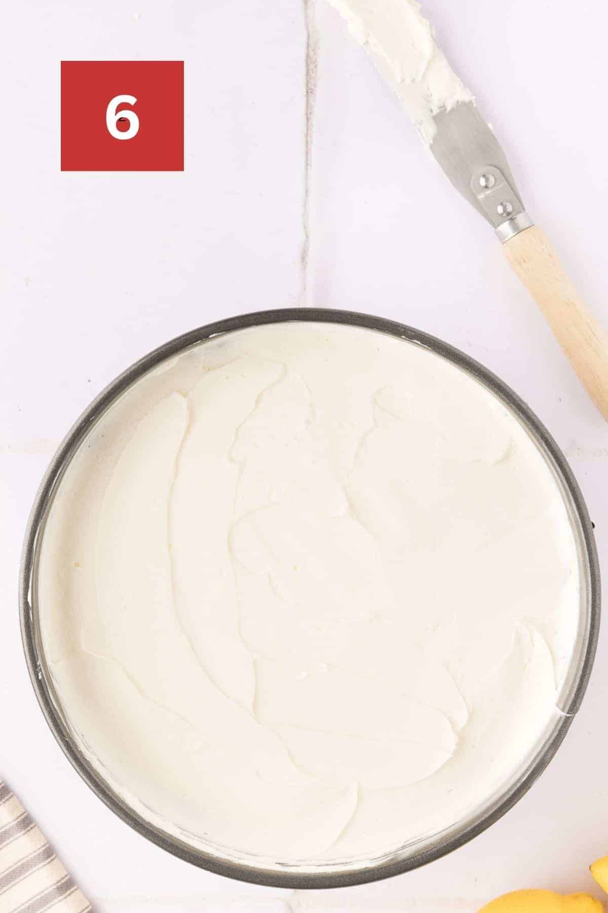 No bake lemon cheesecake in a spring form pan with the cheesecake filling spread evenly on top. It sits on a white tile background with a spatula off to the side. In the upper left corner is a dark red square with a white '6' in the center.