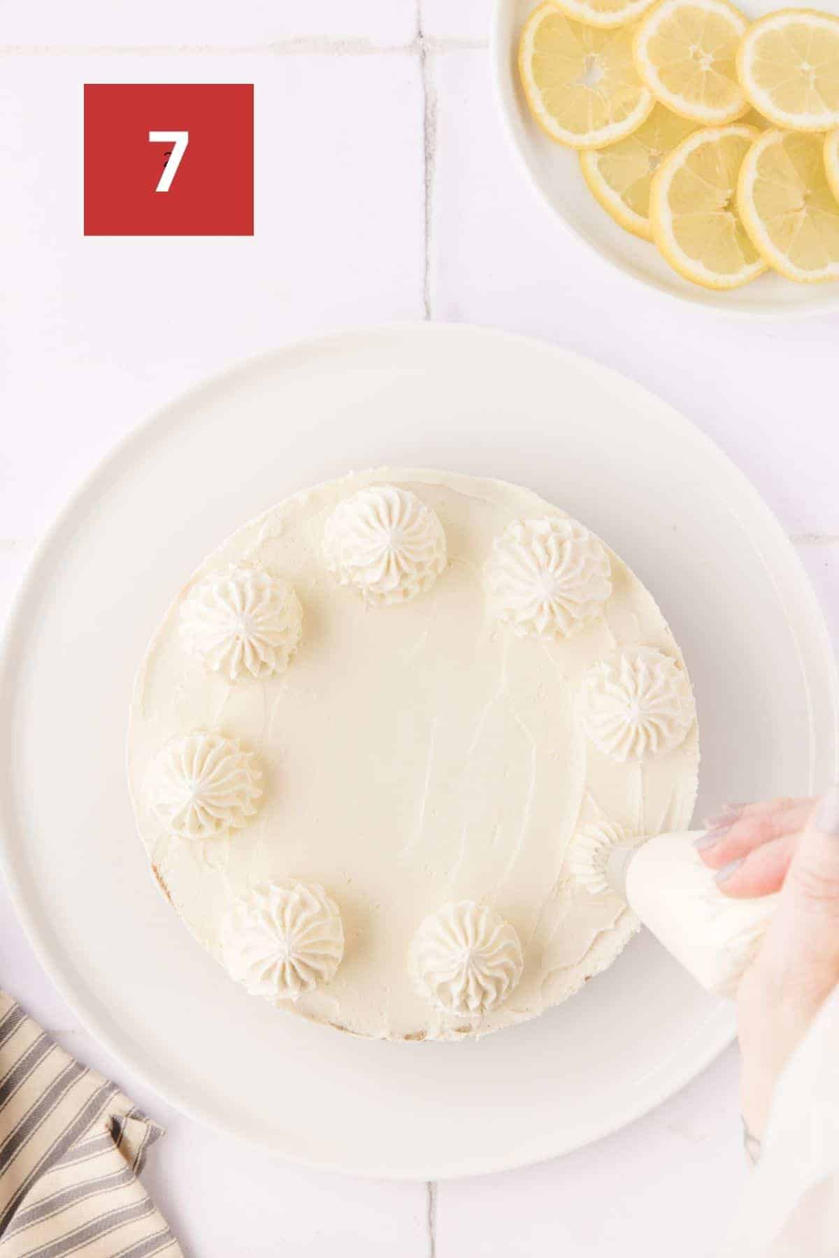 A no bake lemon cheesecake with whip cream being piped on top. The lemon cheesecake sits on a white platter on a white tile background. In the upper left corner is a dark red square with a white '7' in the center.