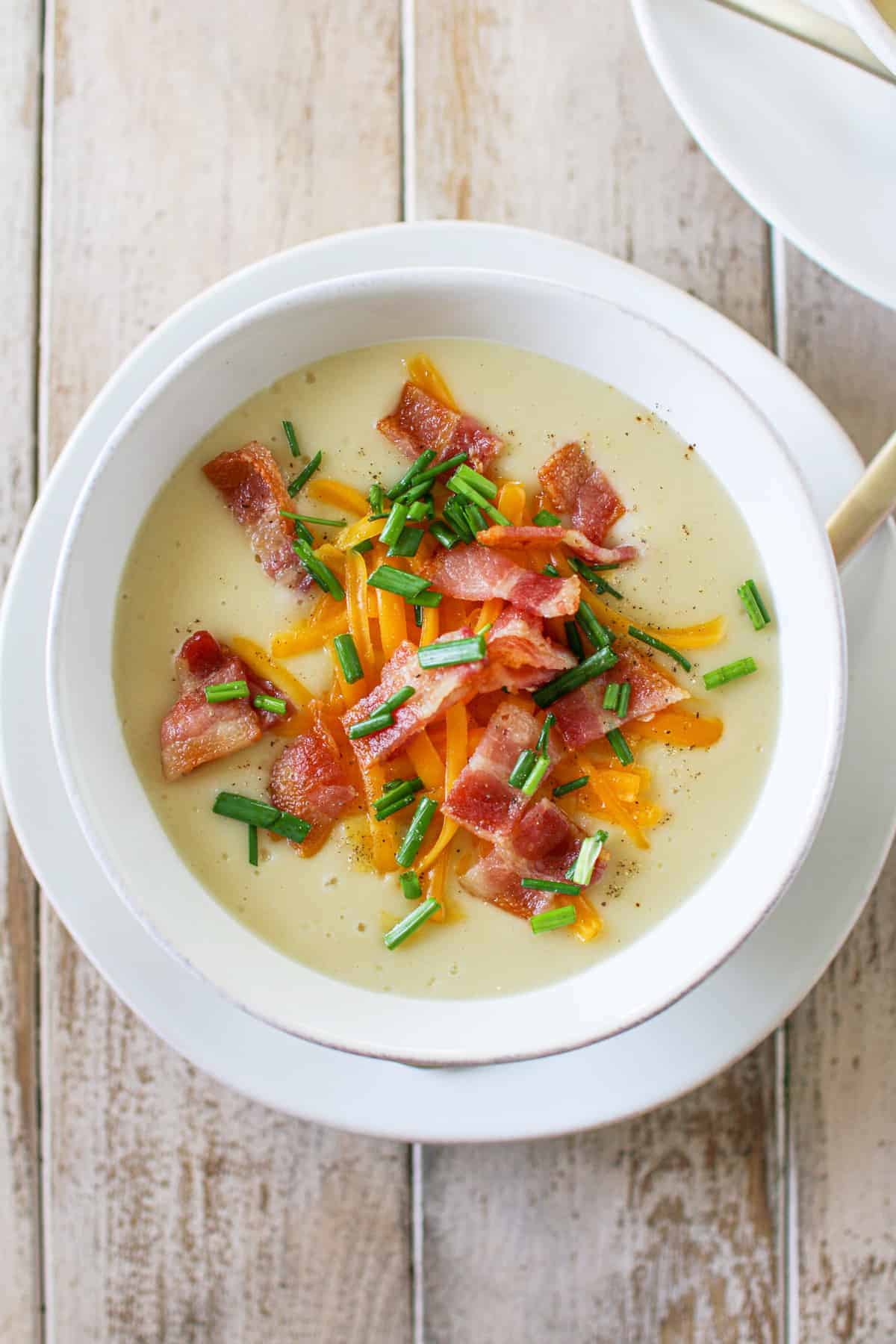 Overhead of a bowl of Potato Cauliflower Soup with thick cut shredded cheddar cheese, bacon, chives and pepper on top. The soup is on a brown wood plank table.