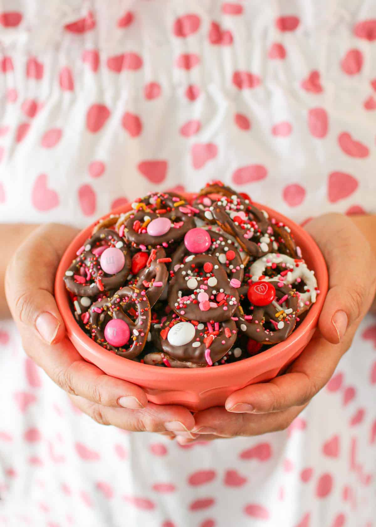Valentine's Day Chocolate Covered Pretzels in a pink bowl being held by two tan hands. The person is wearing a white shirt with pink pots.