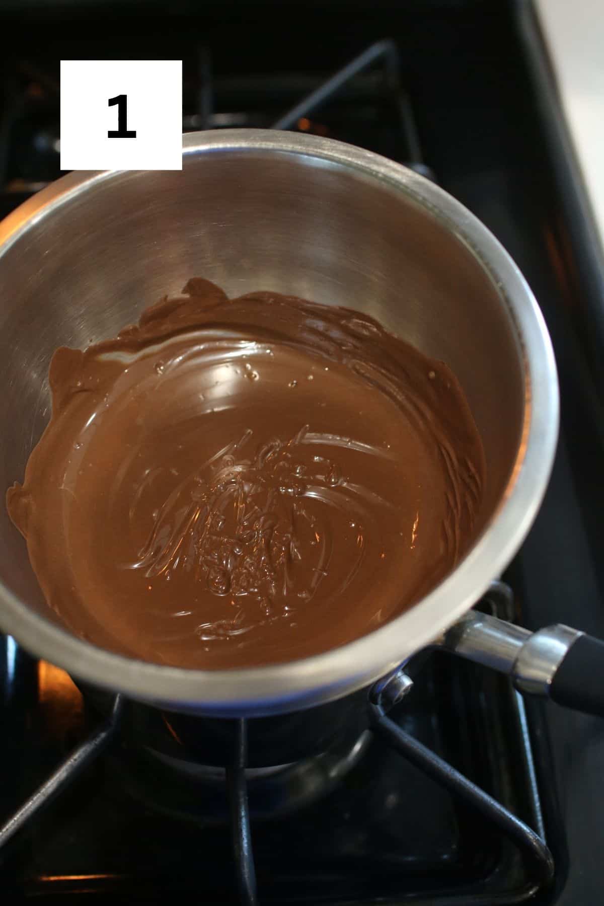 A small metal bowl set up on a double broiler on the stove with melted chocolate. In the upper left corner is a black square with a white '1' in the center.