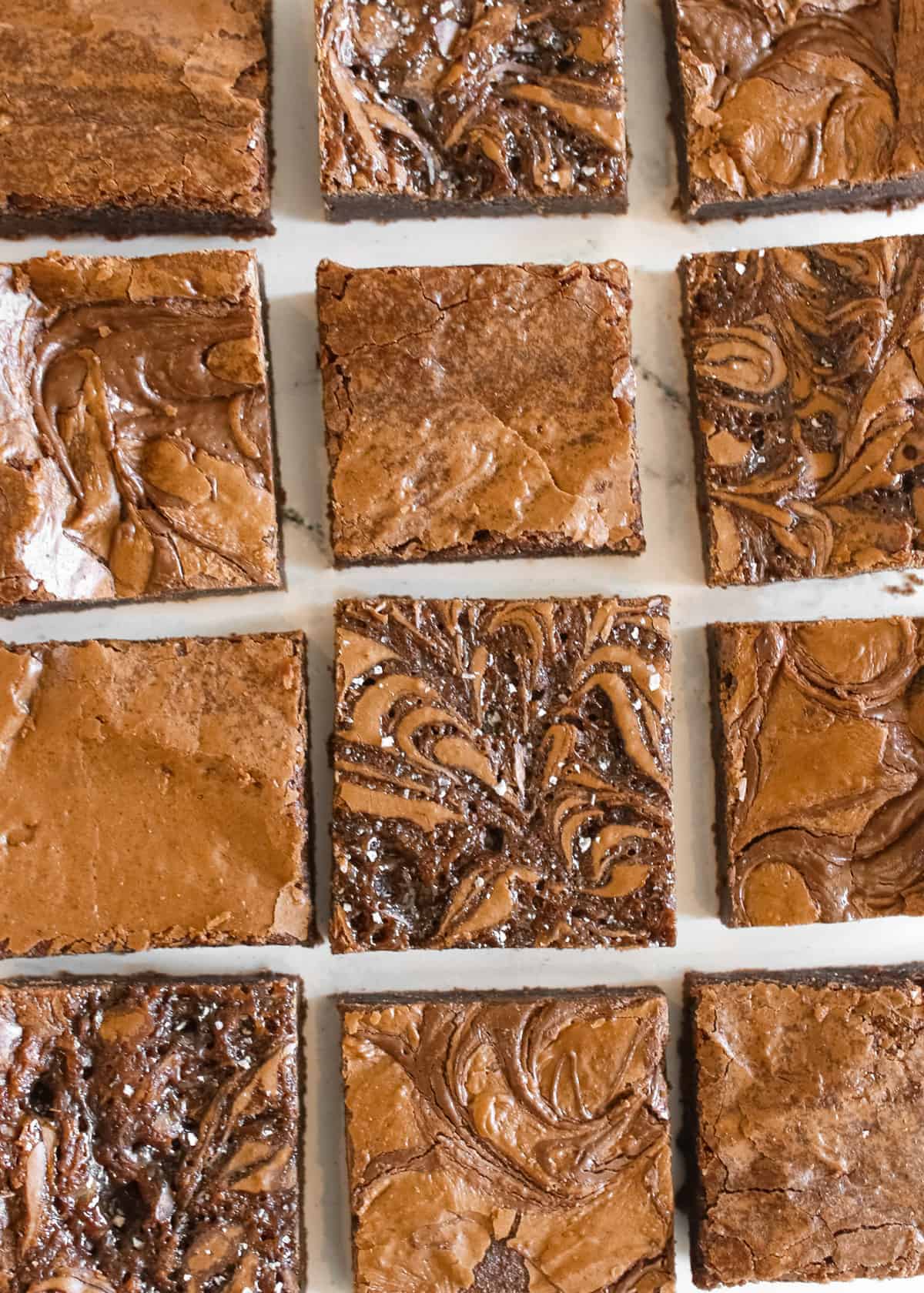 High Altitude Brownies in rows and columns (3x4) with a little space in between each square on a white marble background. 3 variations of brownies include plain, salted caramel and nutella.