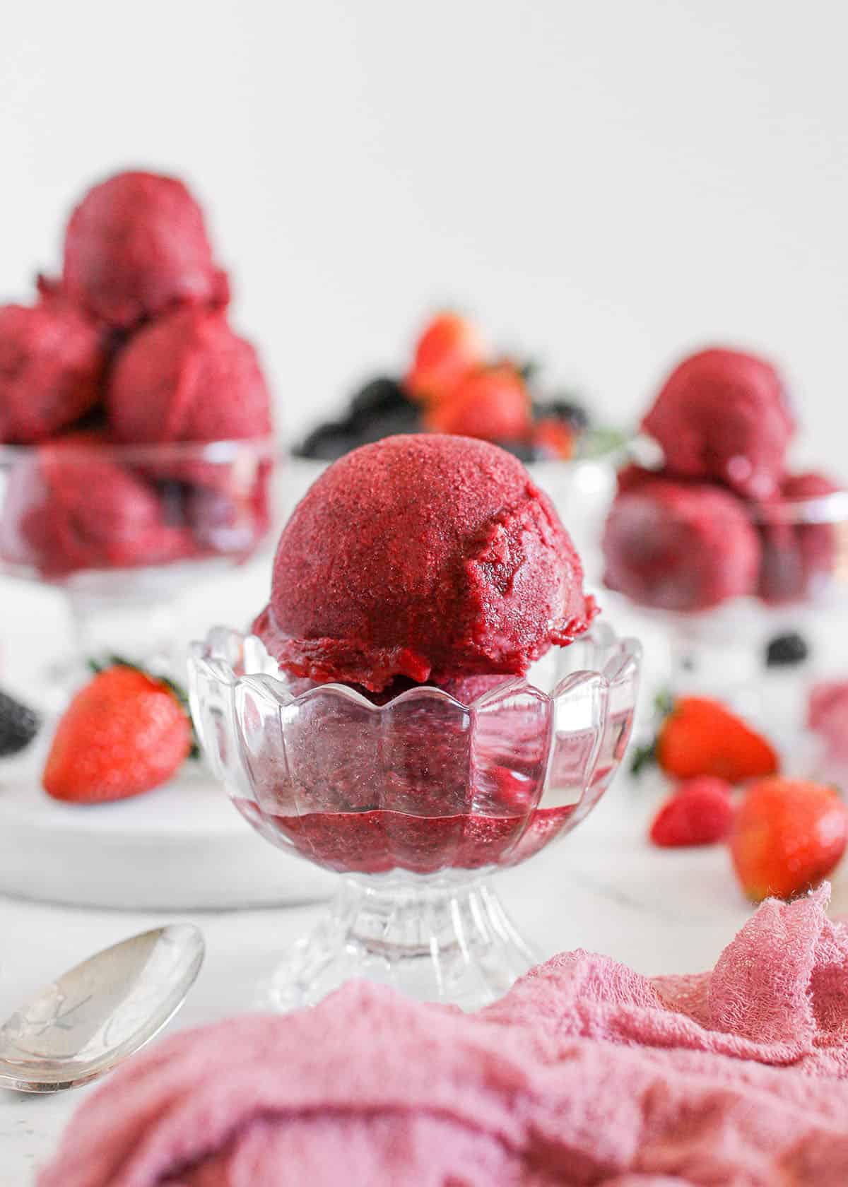 2 scoops of mixed berry sorbet in a glass ice cream bowl. More bowls of sorbet sit in the background along with scattered fresh berries and a mauve gauze sits in front of the ice cream. Everything sits on a white marble table with a white background.