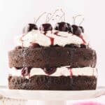 A white scalloped cake stand with a baked black forest cake with layers of whipped cream and cherry compote with fresh cherries on top with cherry syrup drizzled down.