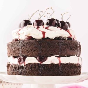 A white scalloped cake stand with a baked black forest cake with layers of whipped cream and cherry compote with fresh cherries on top with cherry syrup drizzled down.