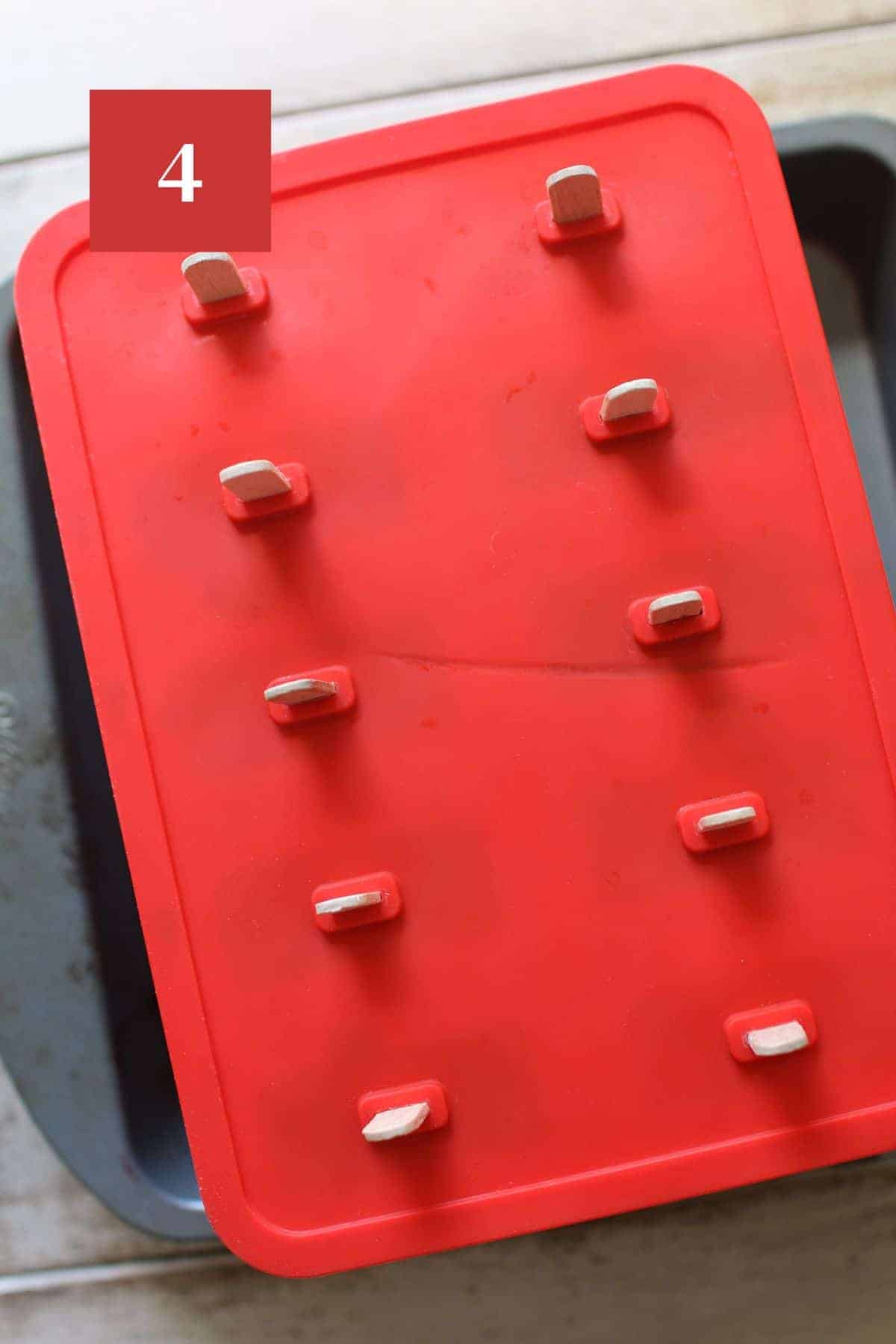 A red popsicle mold in a 8x8 square metal baking pan on a wood plank background. In the upper left corner is a dark red square with a white '4' in the center.