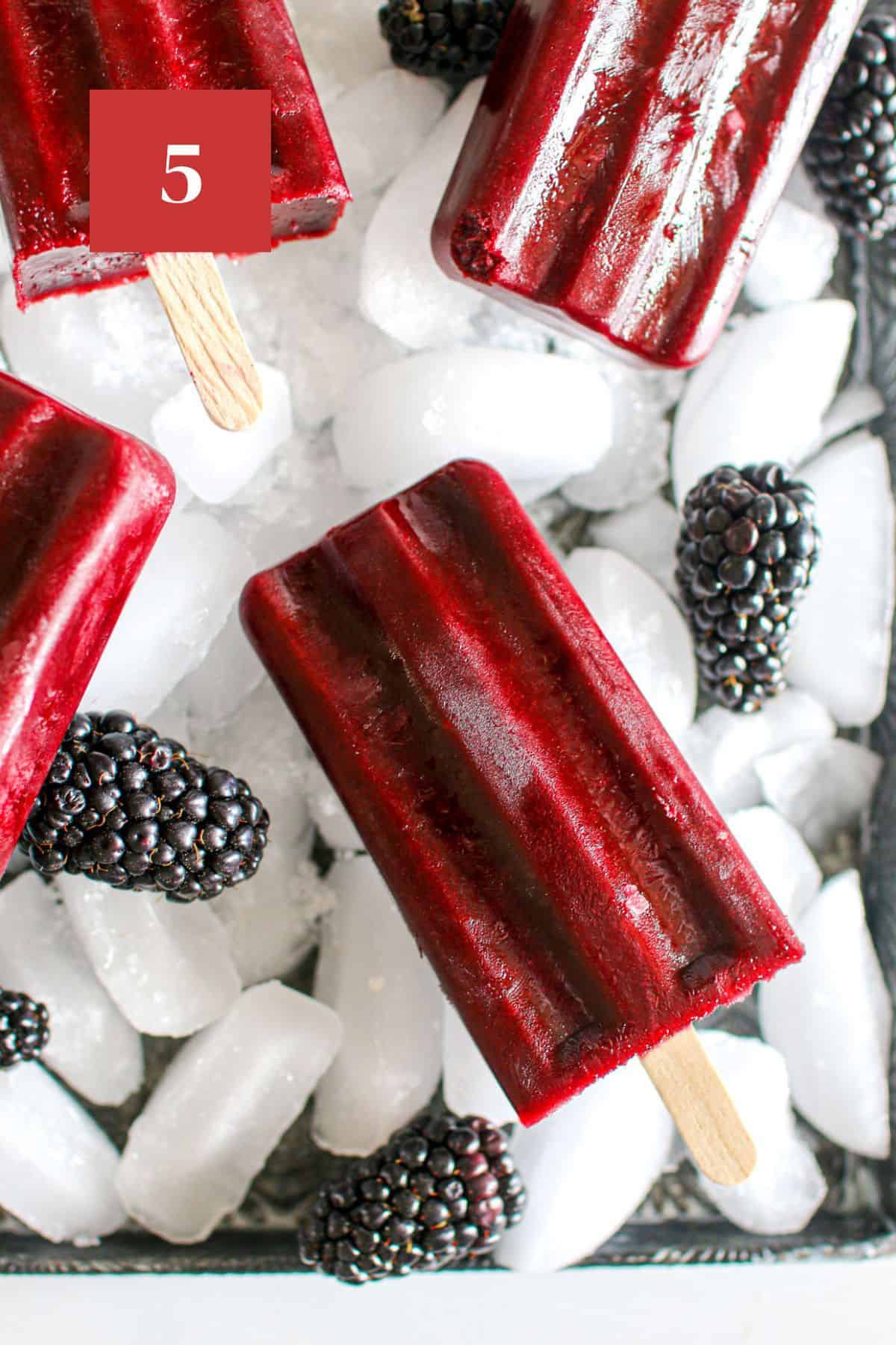 Multiple blackberry popsicles on crushed ice on a baking tray with fresh blackberries surrounding the ice. In the upper left corner is a dark red square with a white '5' in the center.