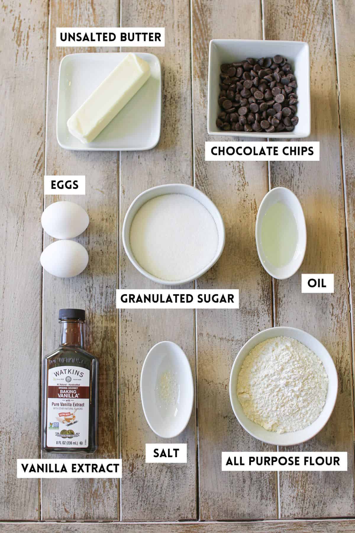 High Altitude Brownie ingredients on a wood plan background. Each ingredient is by items of in a separate vessel. Each is labeled with a white rectangle with black text in all caps.