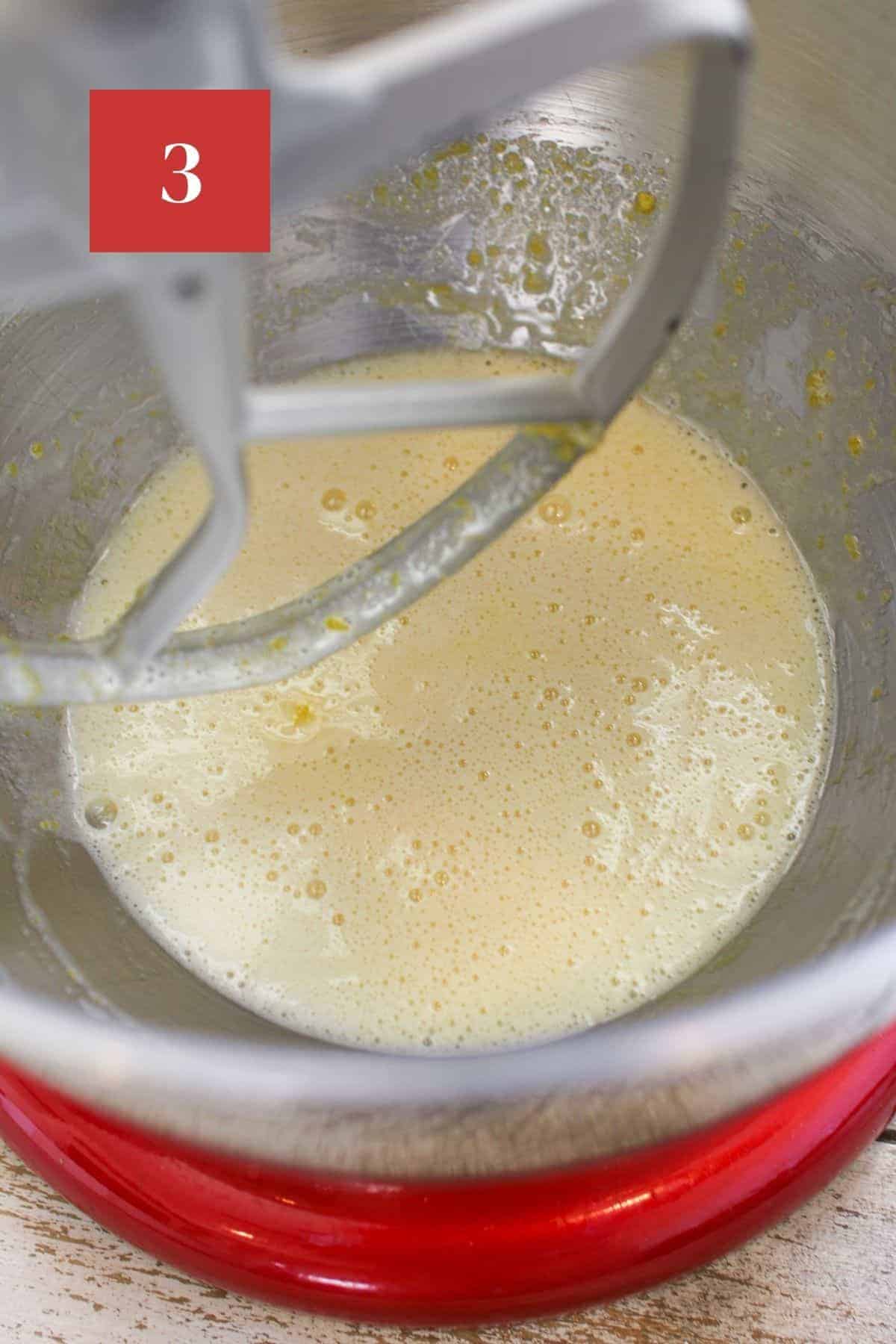 A red stand mixer with eggs, sugar and vanilla being beaten until foamy and a pale yellow.  In the upper left corner is a dark red square with a white '3' in the center.