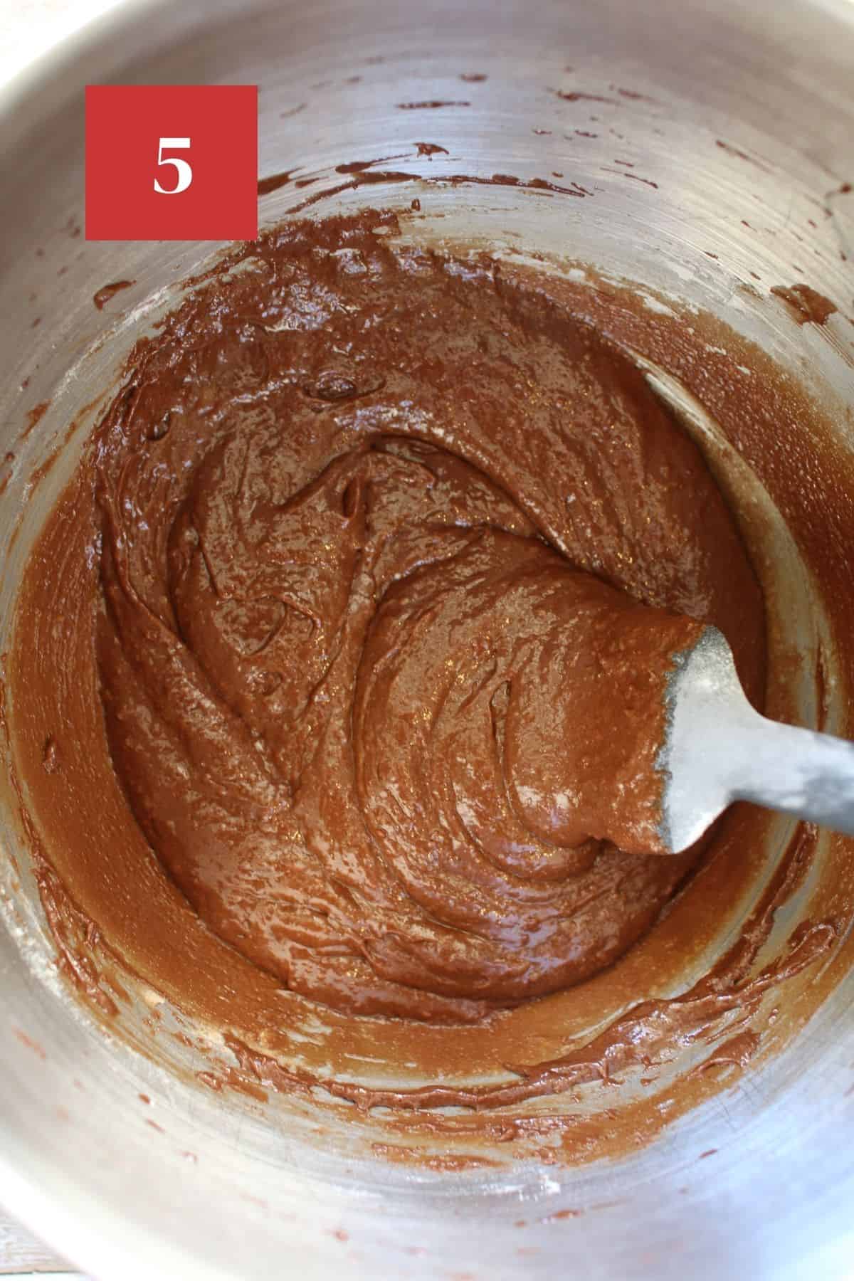 Brownie batter in a large metal mixing bowl with a grey spatula.  In the upper left corner is a dark red square with a white '5' in the center.