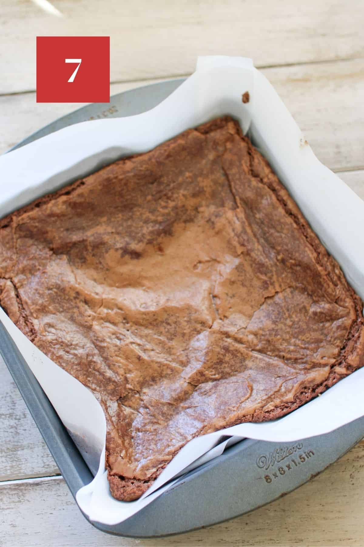 High Altitude brownies baked in a 8x8 baking pan lined with white parchment paper. The pan sits on a wood plank background.  In the upper left corner is a dark red square with a white '7' in the center.