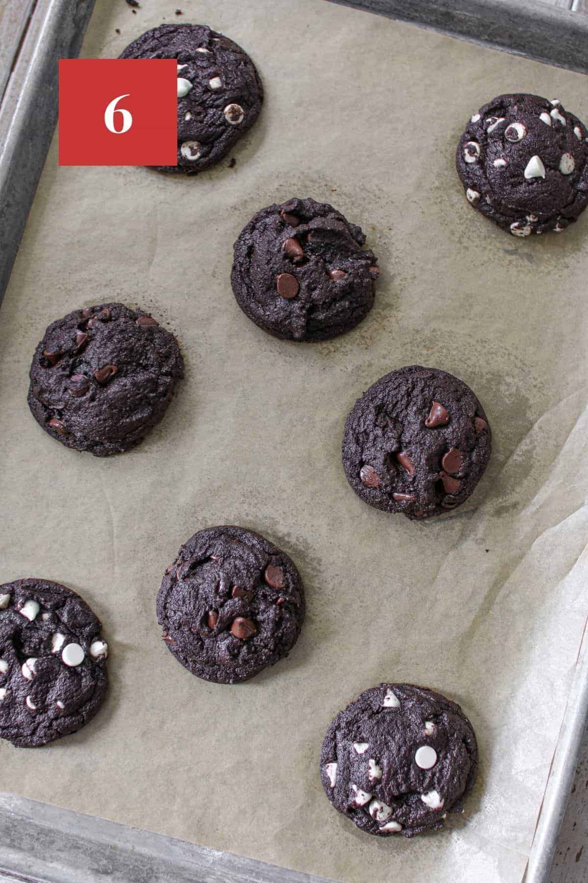 A baking sheet pan with 8 baked double chocolate cookies on the sheet spread out. 4 cookies with white chocolate chips and 4 cookies with semi-sweet chocolate chips. The baking sheet sits on a wood plank background.  In the upper left corner is a dark red square with a white '6' in the center.