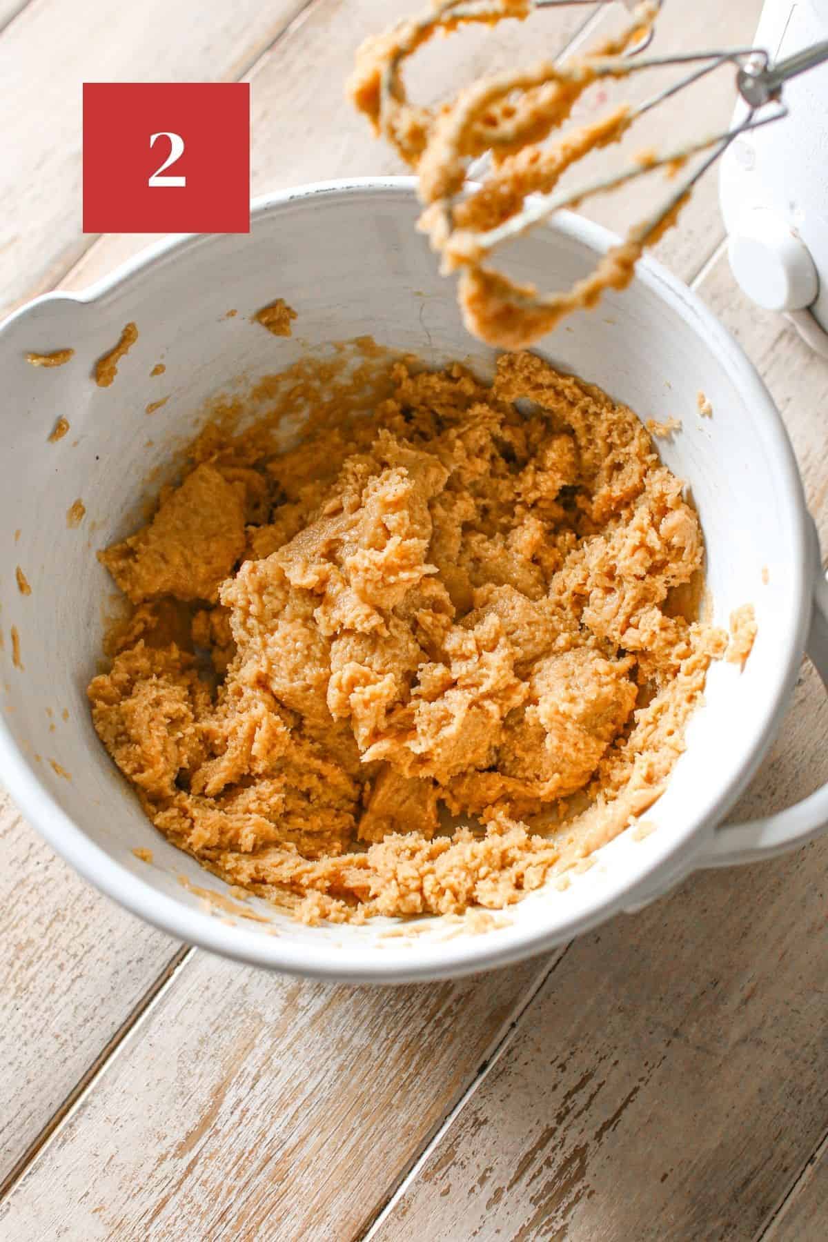 A mixing bowl with peanut butter mix with a hand mixer off to the side on a wood plank background. In the upper left corner is a dark red square with a white '2' in the center.