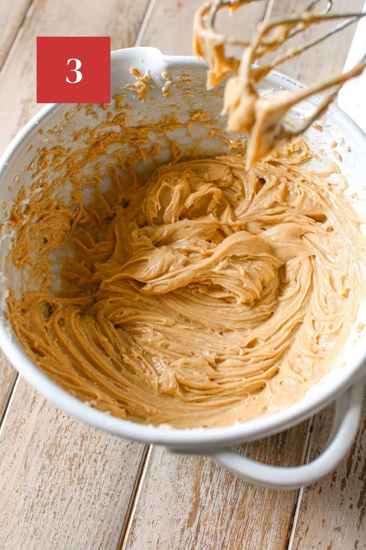 Creamy peanut butter whipped in a mixing bowl with a hand mixer off to the side on a wood plank background. In the upper left corner is a dark red square with a white '3' in the center.