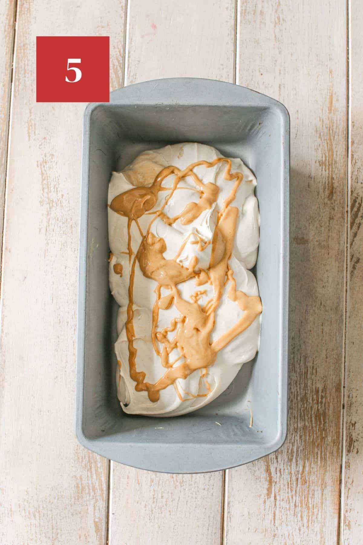 ⅓ of the no churn peanut butter ice cream in a metal loaf pan with melted peanut butter on top. The pan sits on a wood plank background. In the upper left corner is a dark red square with a white '5' in the center.