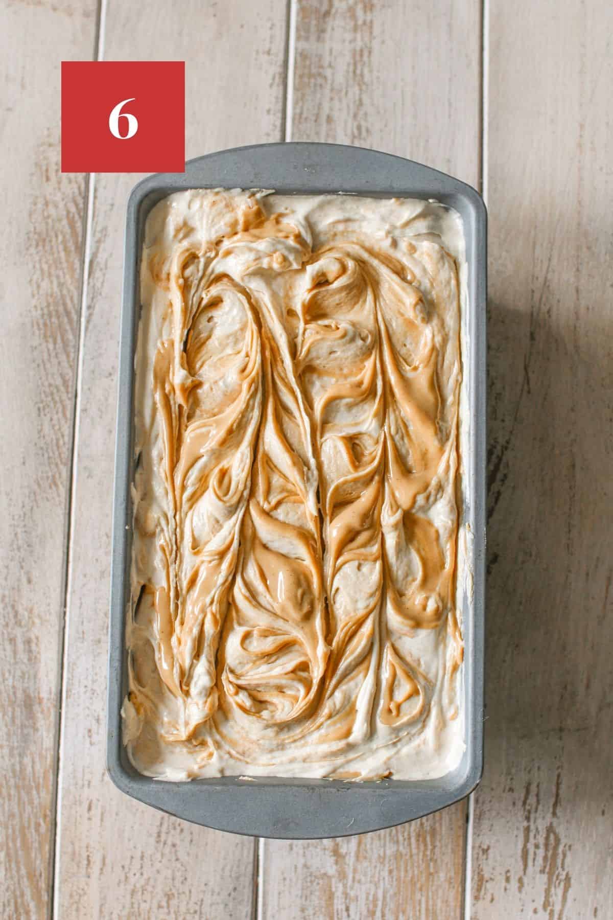 No Churn Peanut butter Ice Cream in a metal loaf pan with peanut butter swirled on top on a wood plank background. In the upper left corner is a dark red square with a white '6' in the center.