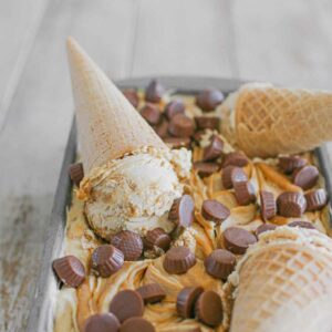 A frozen metal loaf pan with no churn peanut butter ice cream with peanut butter swirled on top with mini peanut butter cups on a light brown wash background. In the loaf pan, there are 3 sugar cones with scooped peanut butter ice creams.