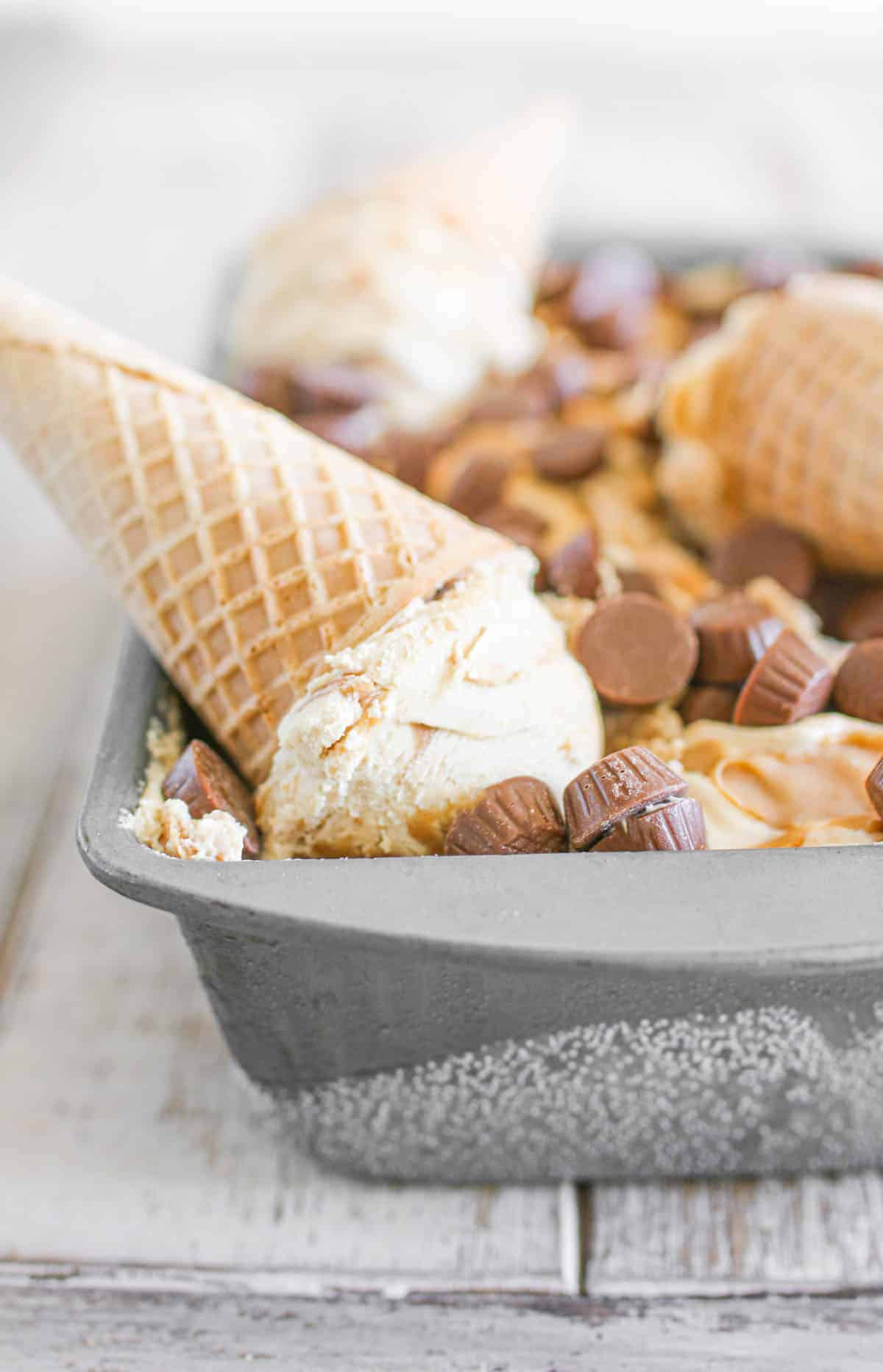 A frozen metal loaf pan with no churn peanut butter ice cream with peanut butter swirled on top with mini peanut butter cups on a light brown wash background. In the loaf pan, there are 3 sugar cones with scooped peanut butter ice creams.