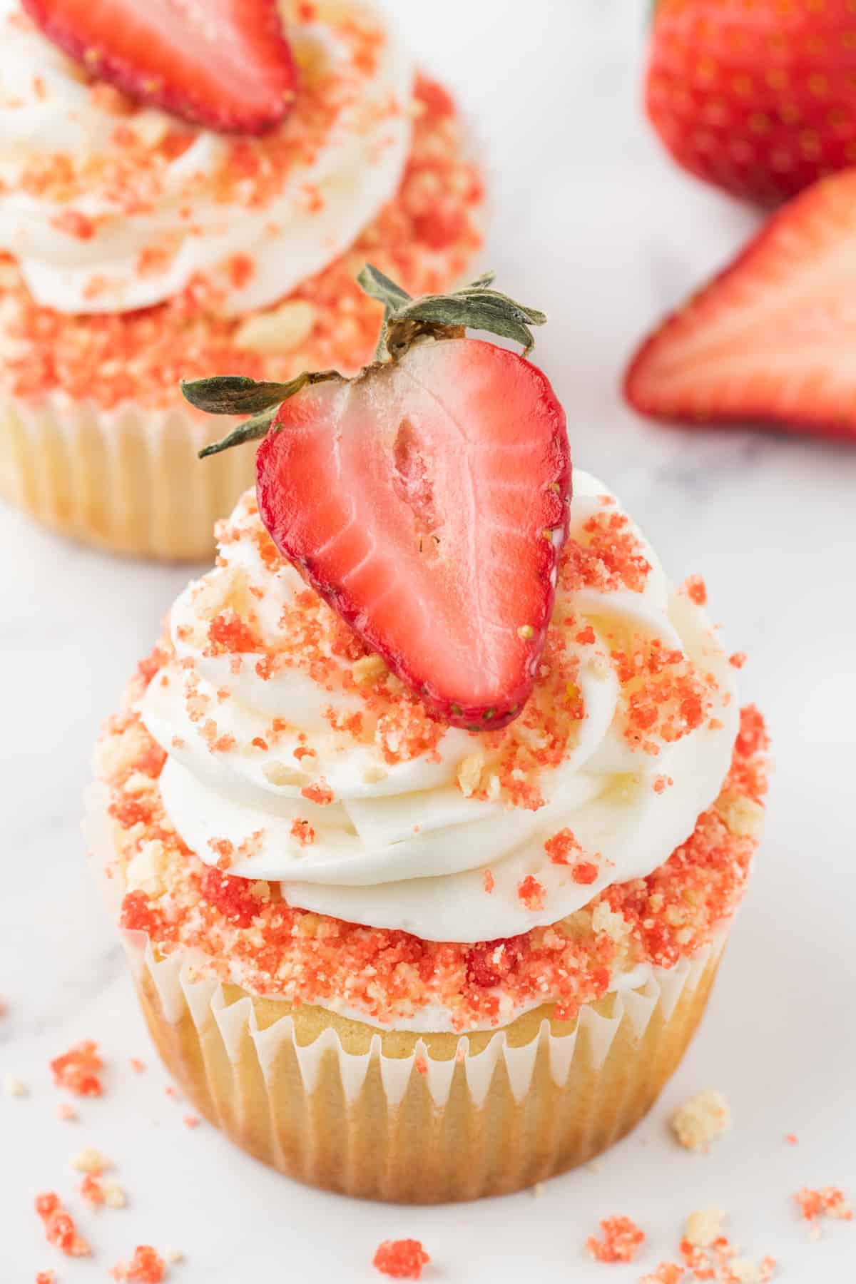 A strawberry crunch cupcake on a white marble background with another cupcake and half strawberry behind it and strawberry crunch topping scattered around the cupcake.