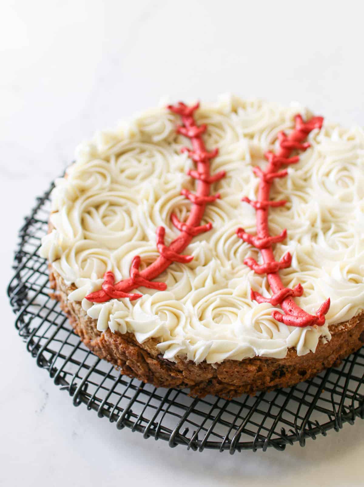 Angled view of Baseball Cookie Cake slightly cut off. The cookie cake sits on a black wire trivet on a white marble background.