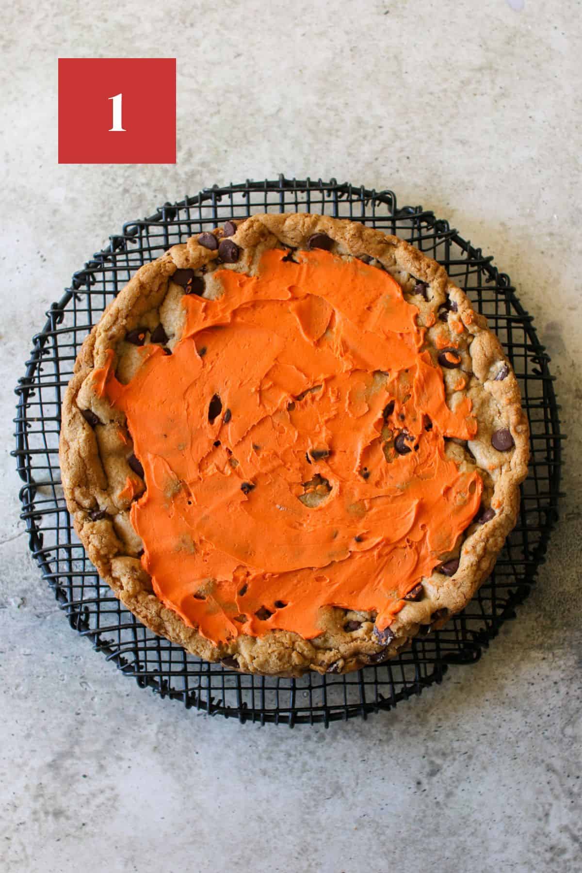 A cookie cake on a black wire trivet on cement background. The cookie cake has a thin layer of orange frosting. In the upper left corner is a dark red square with a white '1' in the center.