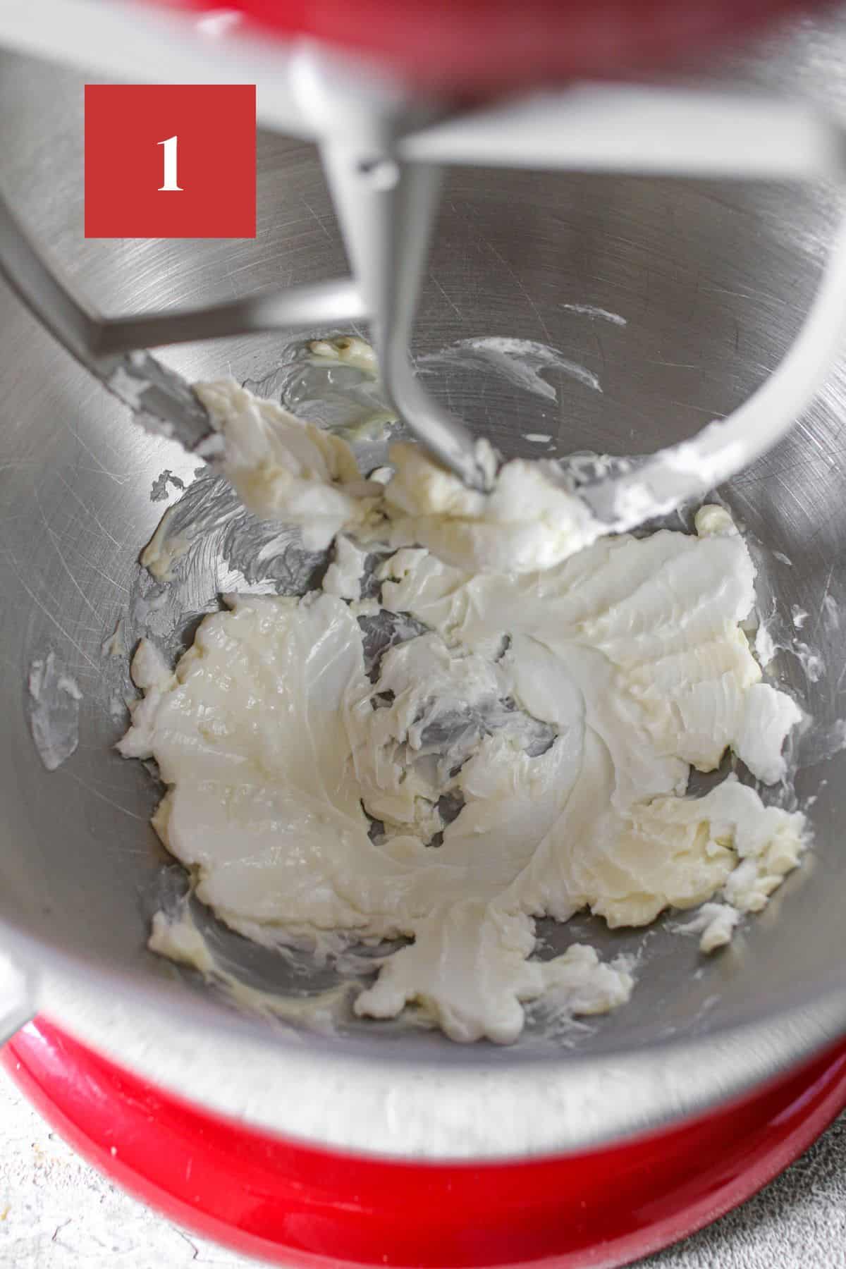 Butter and shortening beaten in a large red stand mixer with a beater attachment. A cookie cake with white rosettes and drop flowers on top. The cookie cake sits on a black wire trivet on a stone background. In the upper left corner is a dark red square with a white '1' in the center.