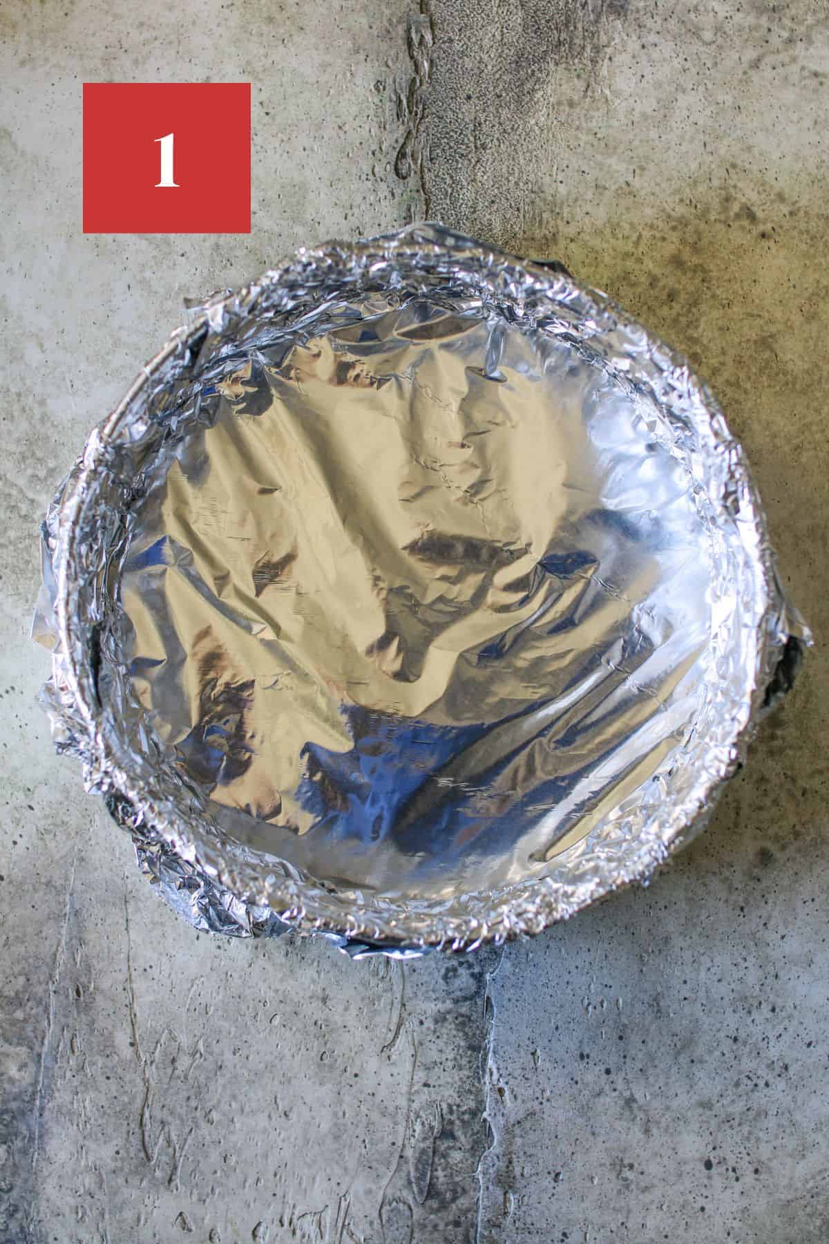 A springform pan wrapped in aluminum foil on a stone background. In the upper left corner is a dark red square with a white '1' in the center.