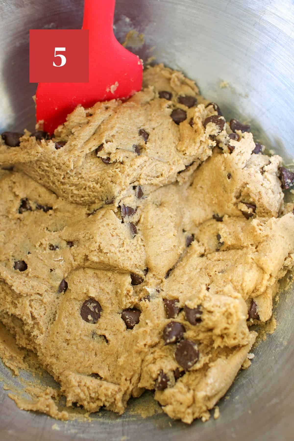 A large steel mixing bowl with chocolate chip cookie cake dough with a red silicone spatula. In the upper left corner is a dark red square with a white '5' in the center.