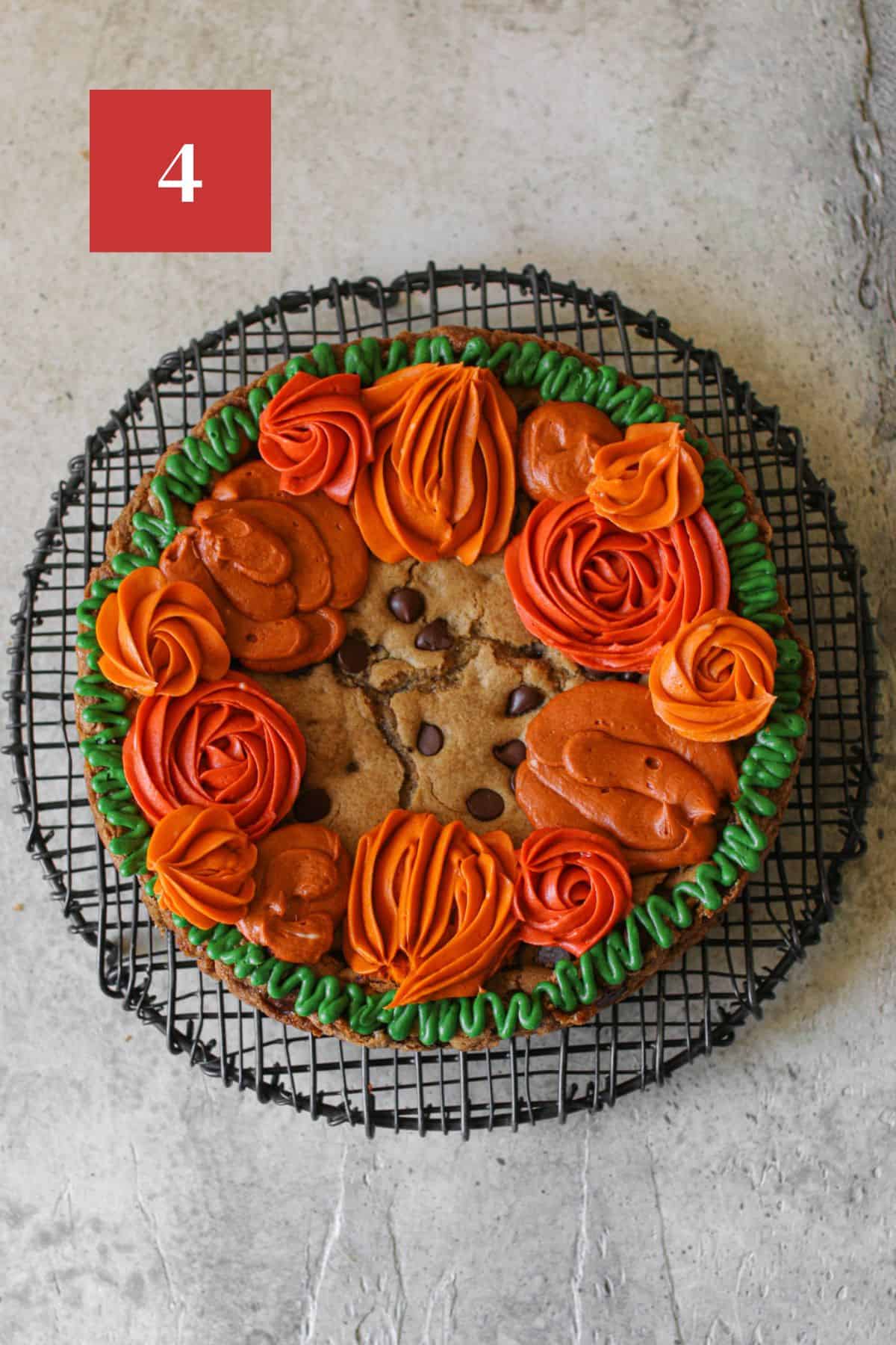 Fall cookie cake with a thin green wavy border and different shades of orange in different sized circles around the edge of the cookie cake. The cookie cake sits on a black wire trivet on a stone background. In the upper left corner is a dark red square with a white '4' in the center.