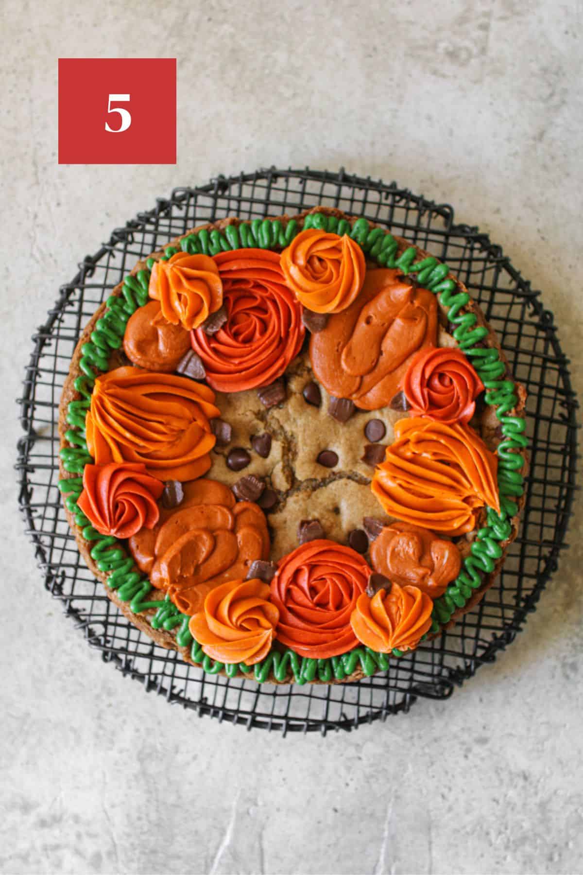 Fall cookie cake with a thin green wavy border and different shades and sizes of orange pumpkins around the edge of the cookie cake. the pumpkins have a small brown stem. The cookie cake sits on a black wire trivet on a stone background. In the upper left corner is a dark red square with a white '5' in the center.