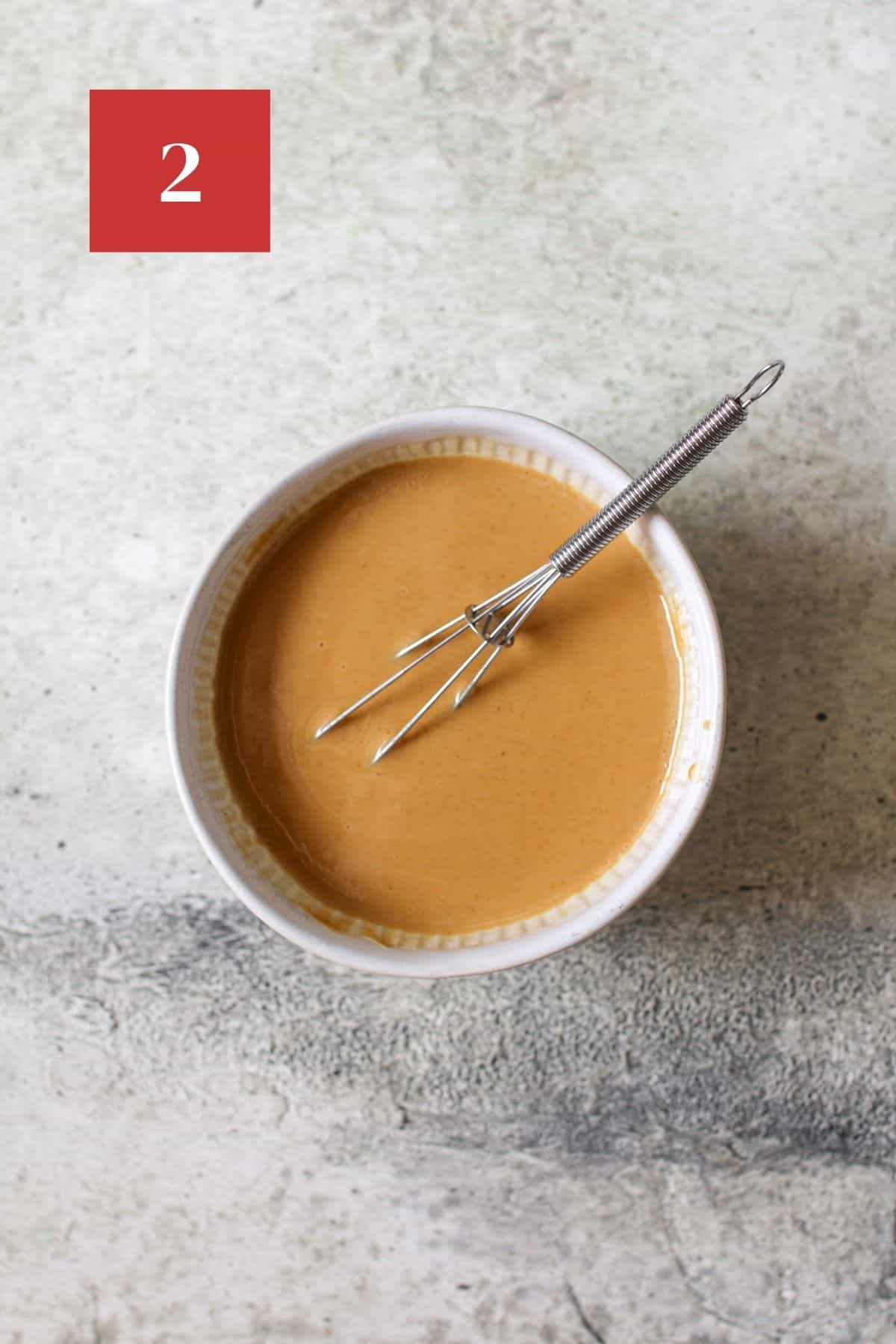 Melted peanut butter and coconut in a small bowl with a mini whisk on a cement background. In the upper left corner is a dark red square with a white '2' in the center.