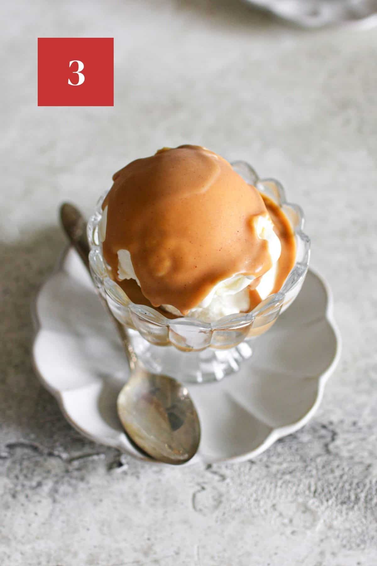 Close up of vanilla ice cream in a clear fluted ice cream glass with hardened peanut butter magic shell. The bowl sits on a small fluted plated with an antique spoon. In the upper left corner is a dark red square with a white '3' in the center.