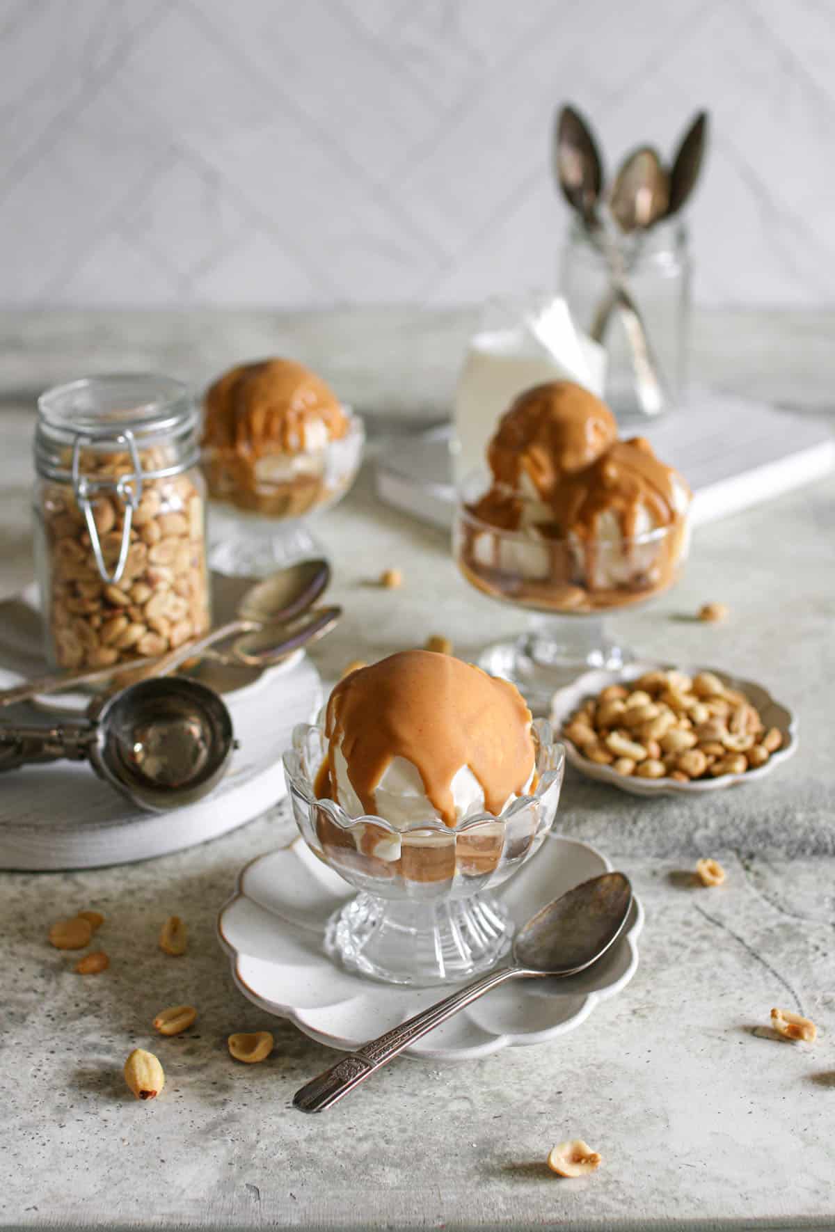 A large scoop of vanilla ice cream with peanut butter magic shell on a scalloped plate with an antique spoon. In the background are white wood boards, more ice cream, peanuts in assorted containers, etc. everything sits on a cement table with a white herringbone background.