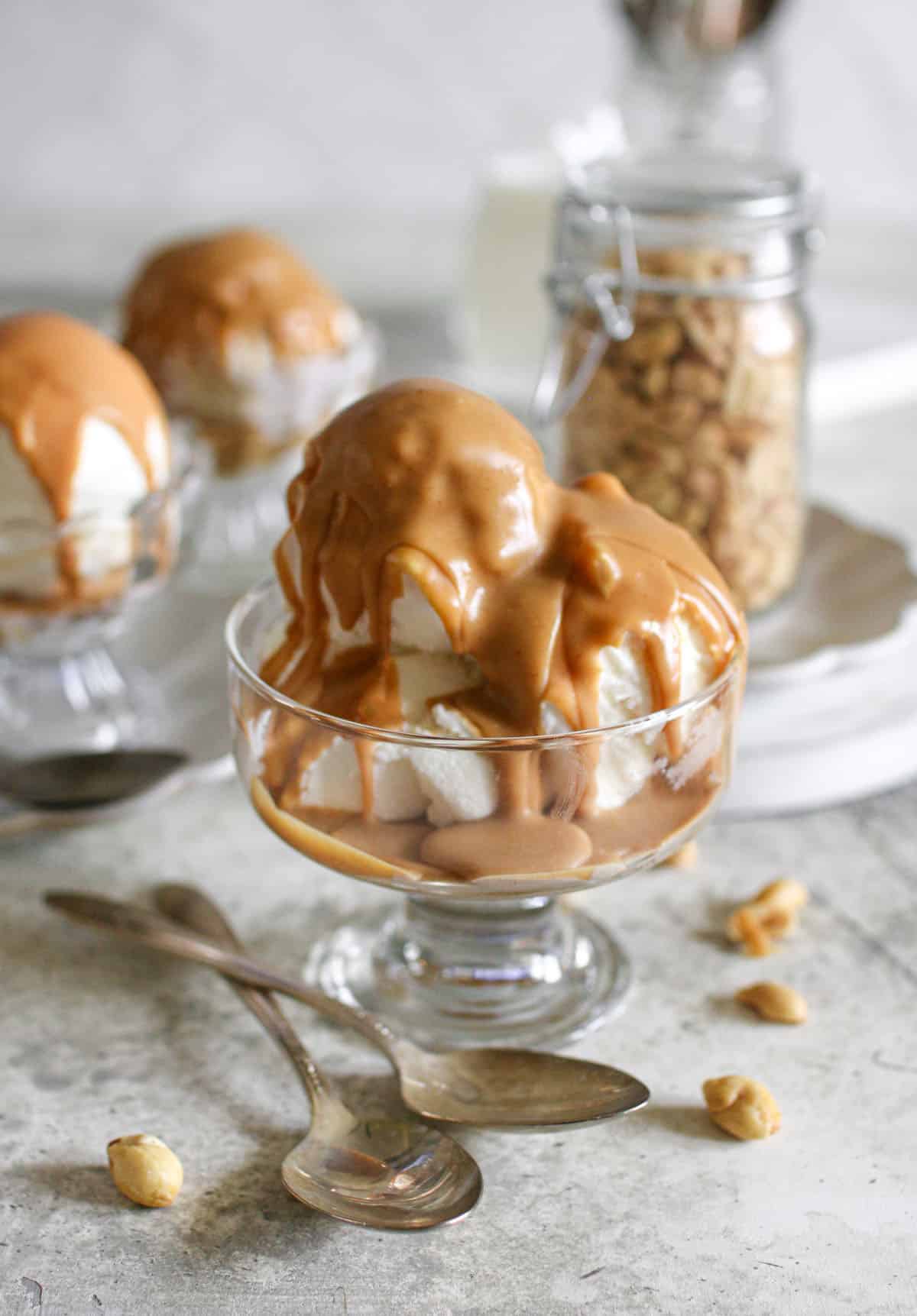Multiple scoops of vanilla ice cream in a clear dish with peanut butter magic shell on top. It sits on a cement background with more ice cream in the back and surrounded by peanuts and antique spoons. 
