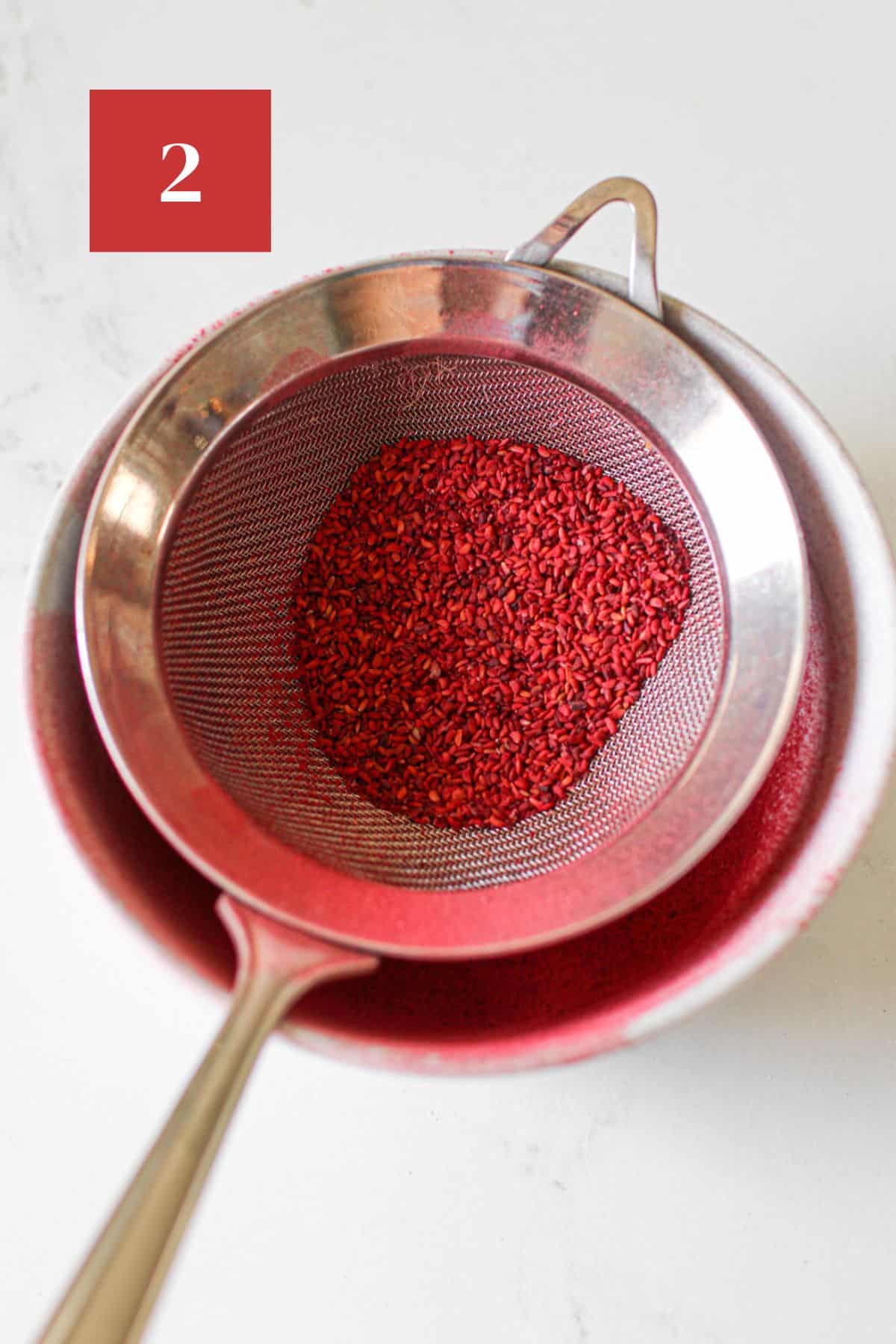  Raspberry freeze dried powder in a small fine mesh strainer with seeds in the strainer in a white bowl on a white table. In the upper left corner is a dark red square with a white '1' in the center.