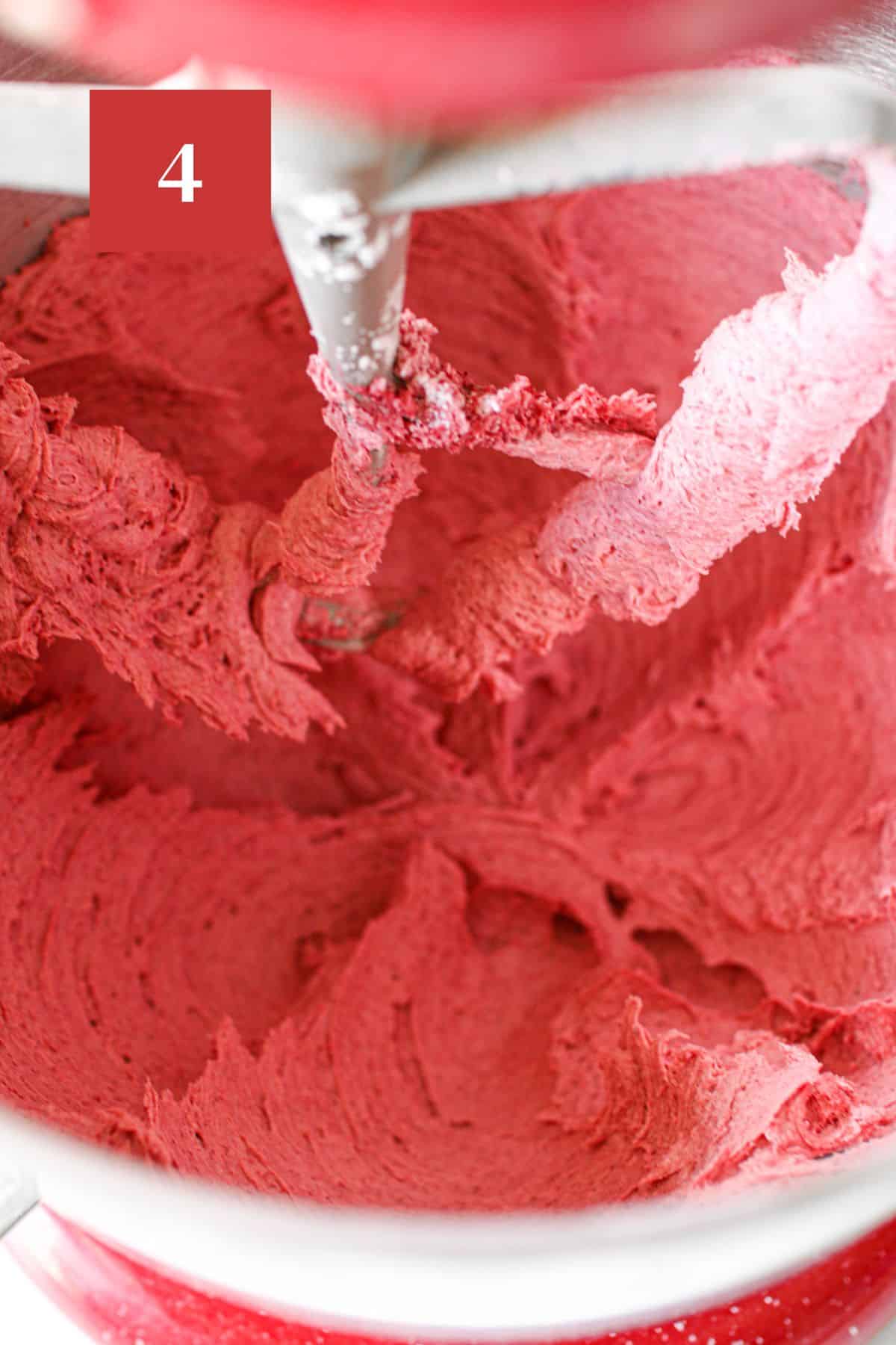 Raspberry buttercream frosting beaten in a large red stand mixer with beater attachment.  In the upper left corner is a dark red square with a white '4' in the center.