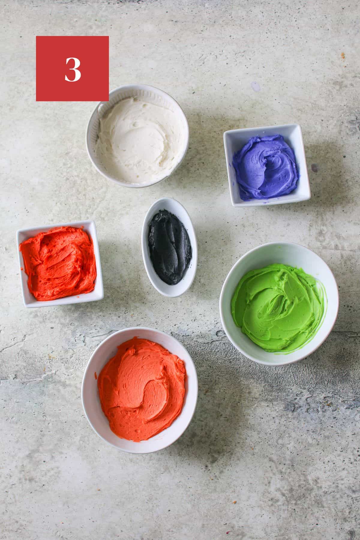 6 white bowls of different sizes sit on a cement background. Each is filled with a different frosting color - white in a circle bowl, violet in a mini square bowl, orange red in a mini square bowl, orange in a small low bowl, lime green in a small low bowl and black in a small oval bowl.  In the upper left corner is a dark red square with a white '3' in the center.