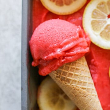 A large scoop of Strawberry Lemonade Sorbet on a sugar cone in a tin with more sorbet and sliced lemons. The tin sits on aw cement background.