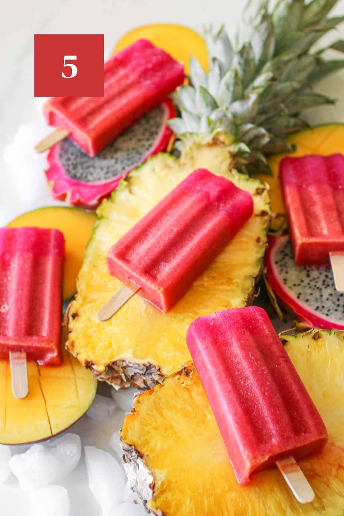 Angled view of Tropical Popsicles on a variety of sliced tropical fruit - pineapple, mango and dragon fruit with crushed ice surrounding the fruit on a white background. In the upper left corner is a dark red square with a white '5' in the center.