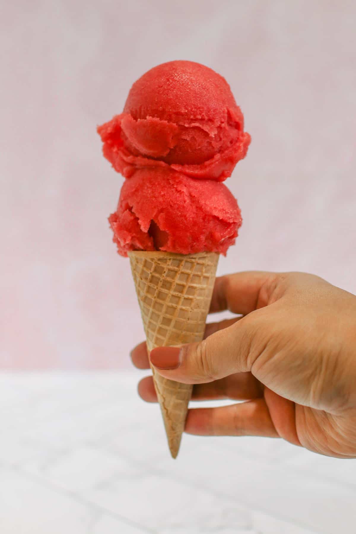 2 scoops of strawberry lemonade sorbet on a sugar cone. A hand is holding the cone. In the background is a pink marble wall.