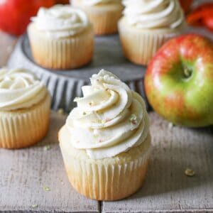 A vanilla cupcake with a white line with apple buttercream stacked tall on top. It has freeze dried apple bits decorating the cupcake. Behind it are more cupcakes and whole apples. Everything sits on a wood plank table.