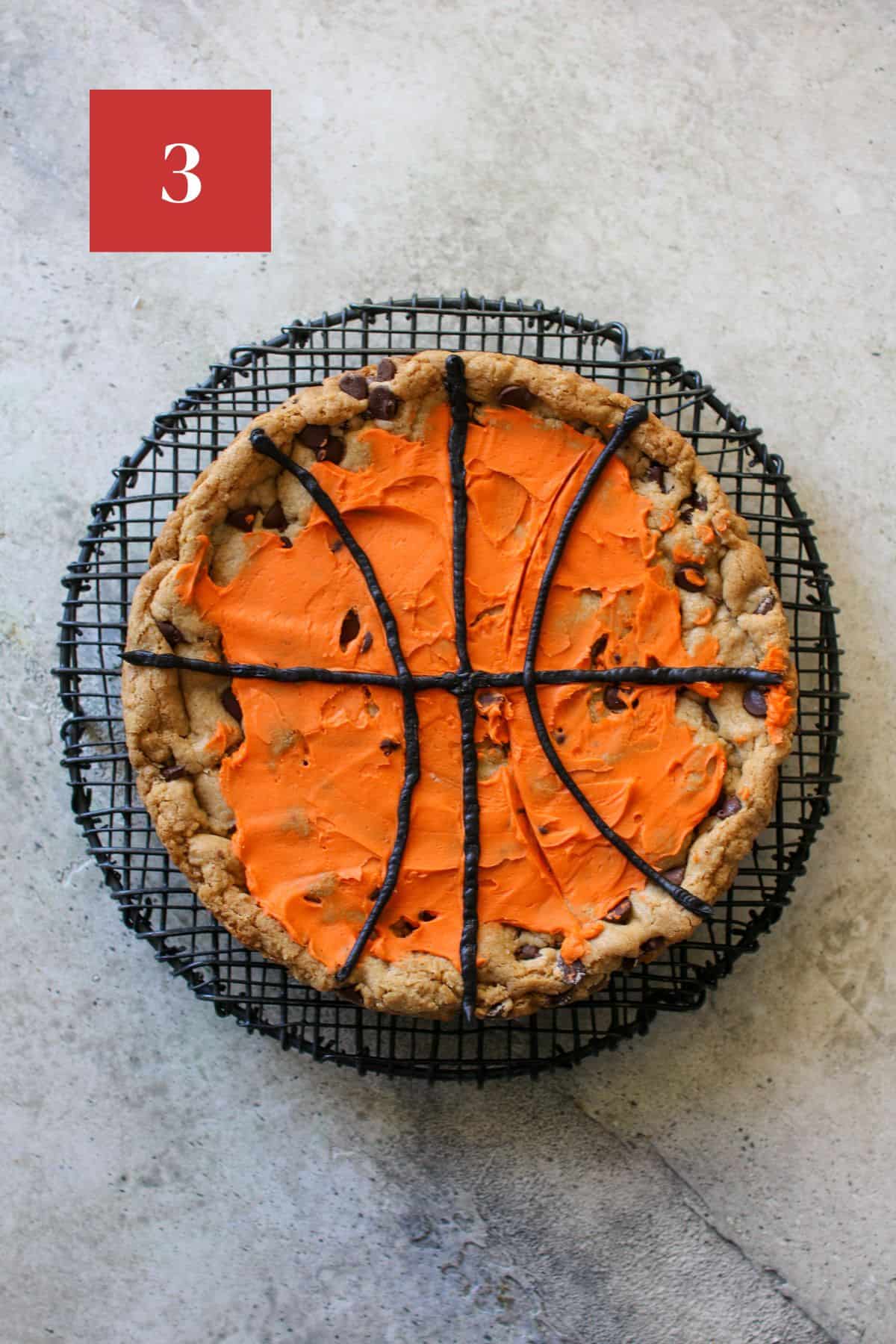 A cookie cake on a black wire trivet on cement background. The cookie cake has a thin layer of orange frosting and basketball lines drawn with a small offset spatula. On top of the orange frosting are thin black basketball lines made of frosting.  In the upper left corner is a dark red square with a white '3' in the center.