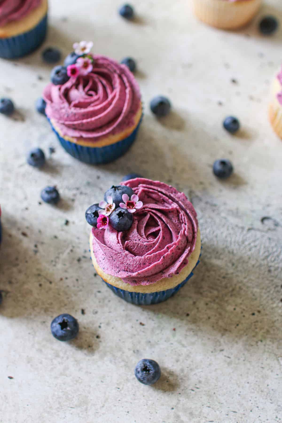 Blueberry buttercream on vanilla cakes lined with a blue cupcake liner. The blueberry buttercream is swirled on top of the cupcake and has fresh blueberries and small pink flowers. The cupcakes sit on a cement background and surrounded by other cupcakes and more fresh blueberries.